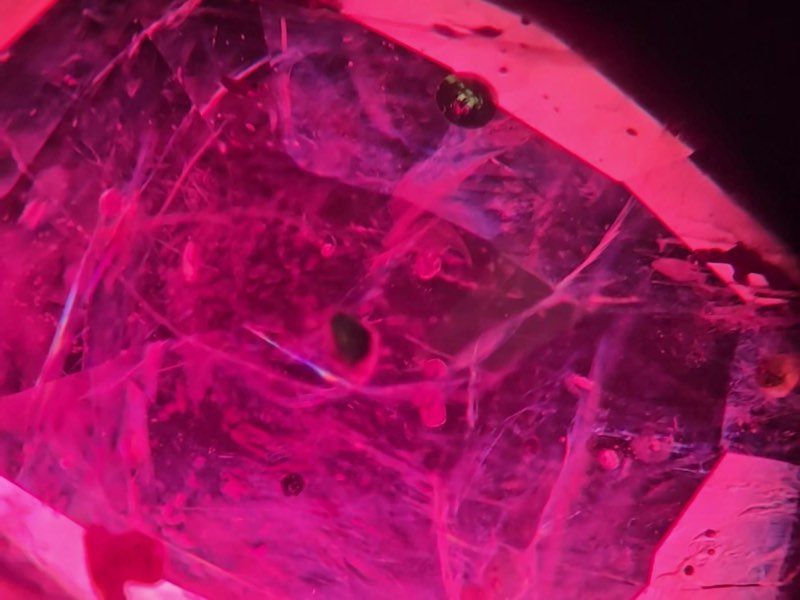 Vue au microscope d’un rubis rempli de verre : un réseau de fines fissures et de petites taches rondes à l’intérieur d’une pierre rouge rosé profond, aux arêtes de facettes d’un rose vif